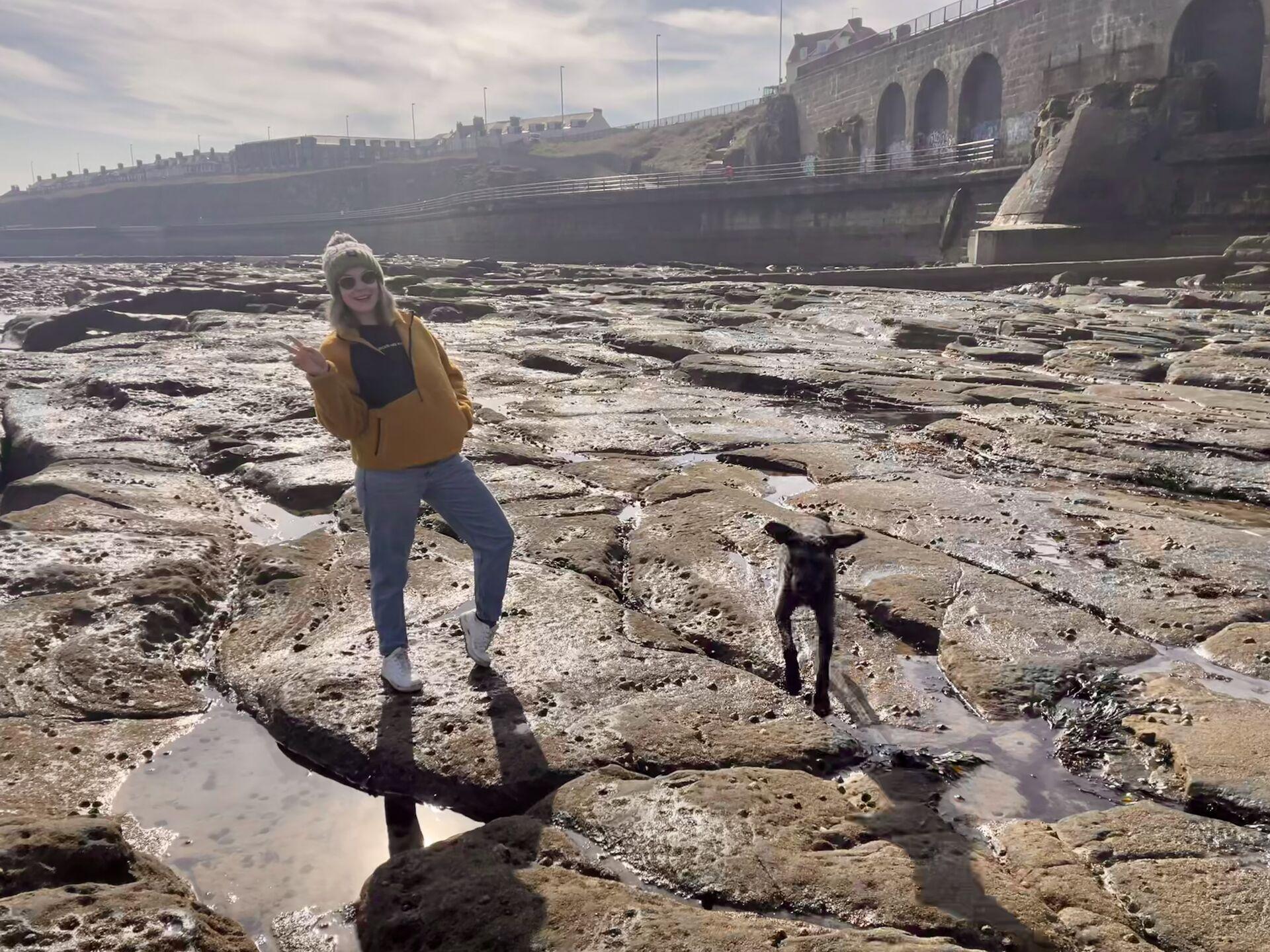 A woman in a yellow jacket and gray pants posing on a rocky shoreline with a black dog nearby, with a bridge and buildings in the background.