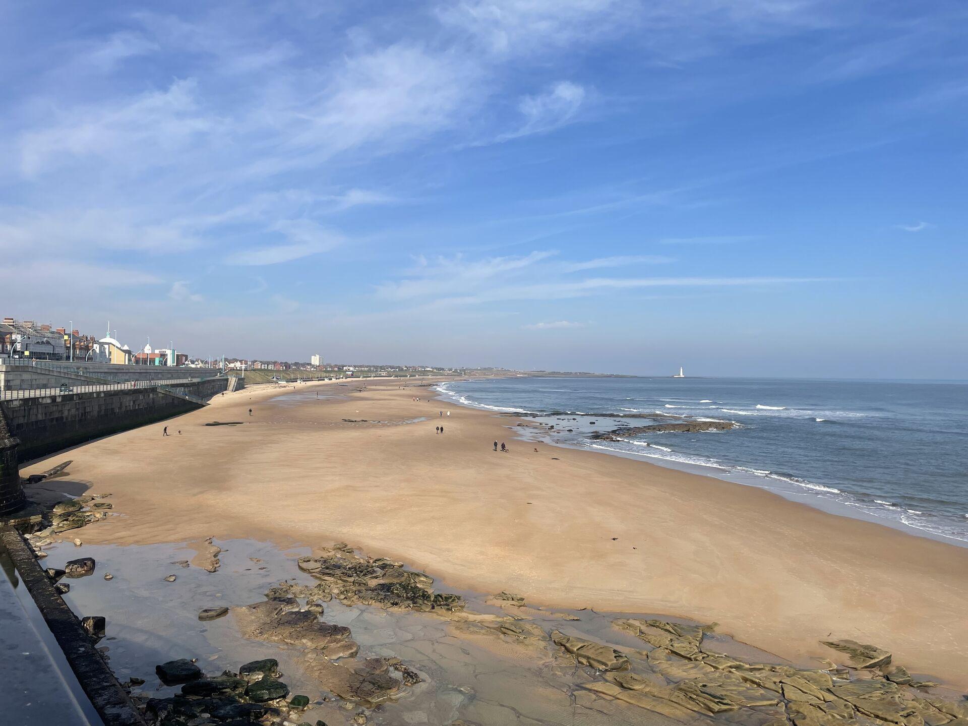 A wide sandy beach with a few people walking, a calm ocean with gentle waves, a rocky shoreline in the foreground, and a promenade with buildings along the coast under a partly cloudy sky.