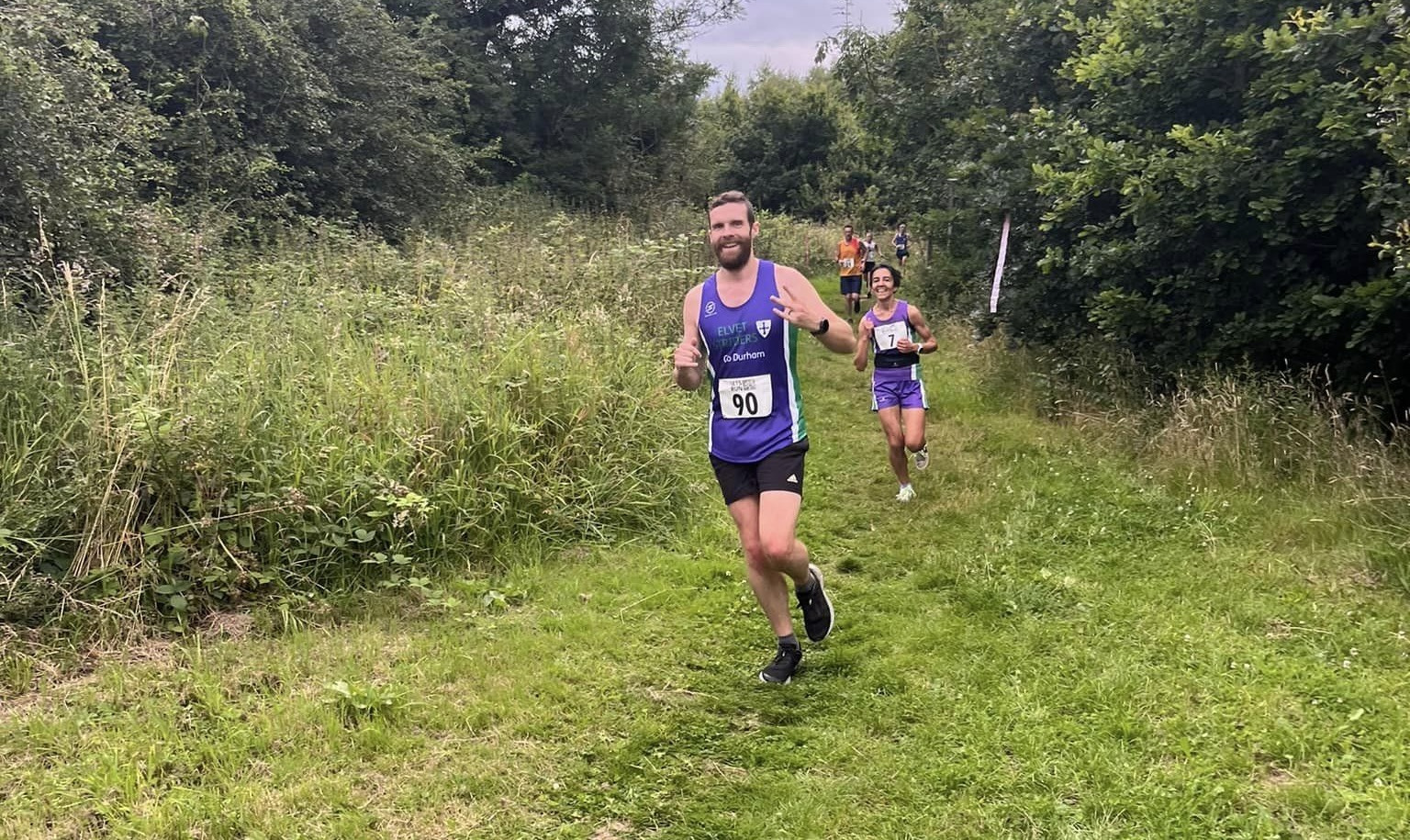 Me, in a purple and green race jersey, running down a grassy path between brambly-looking trees, followed closely by other runners about to pass me