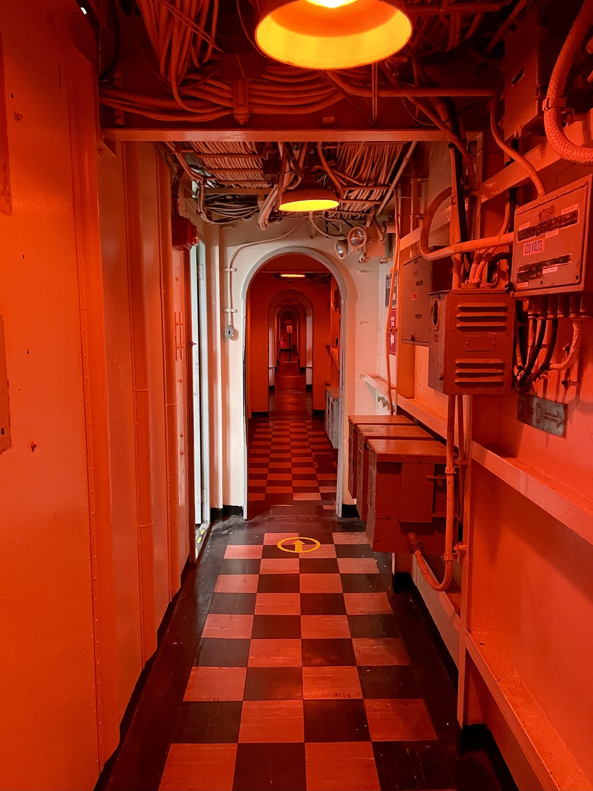 A long hallway on board the USS Yorktown, with red lighting overhead and a checkboard floor and lots of wires running overhead.