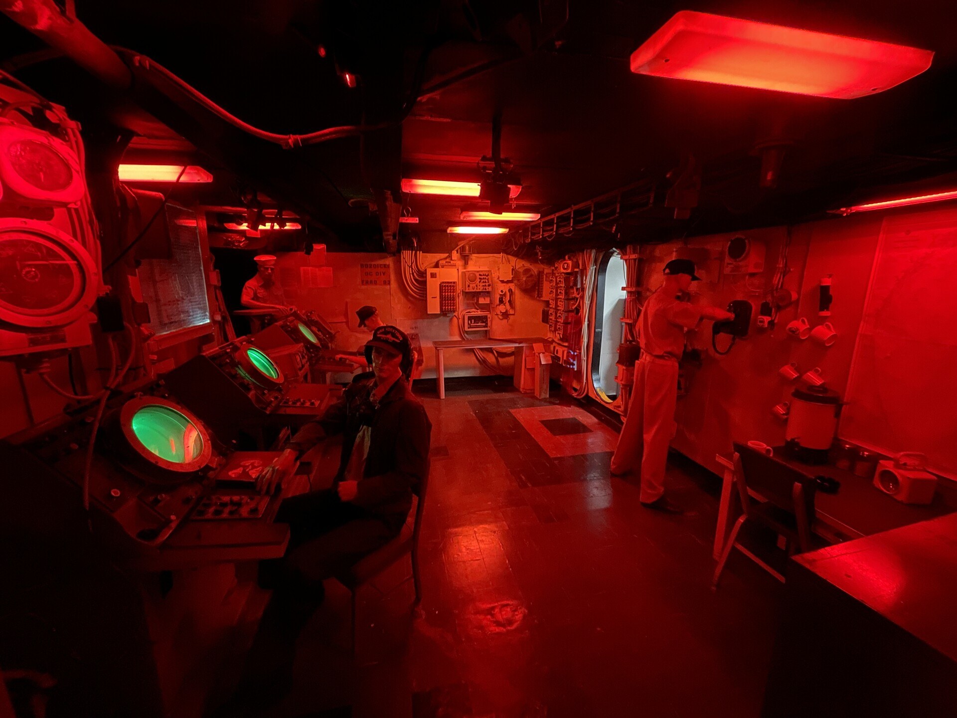 The radar room aboard USS Yorktown, with a few awkward mannequins standing around under the red light. One mannequin is turned towards the viewer as if about to engage in conversation.